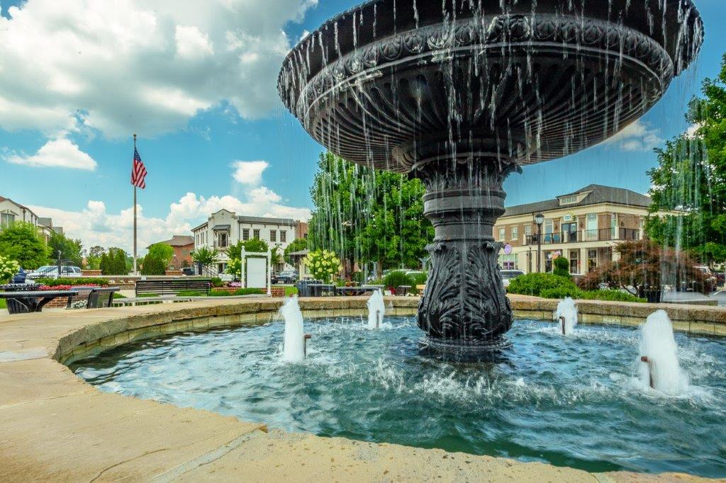 Picture of water fountain in Cambridge square in Ooltewah TN.   Runnging water over the fountain in a circle shape like champaign goblet.  buisnesses made to look more like homes in the back ground and US Flag
