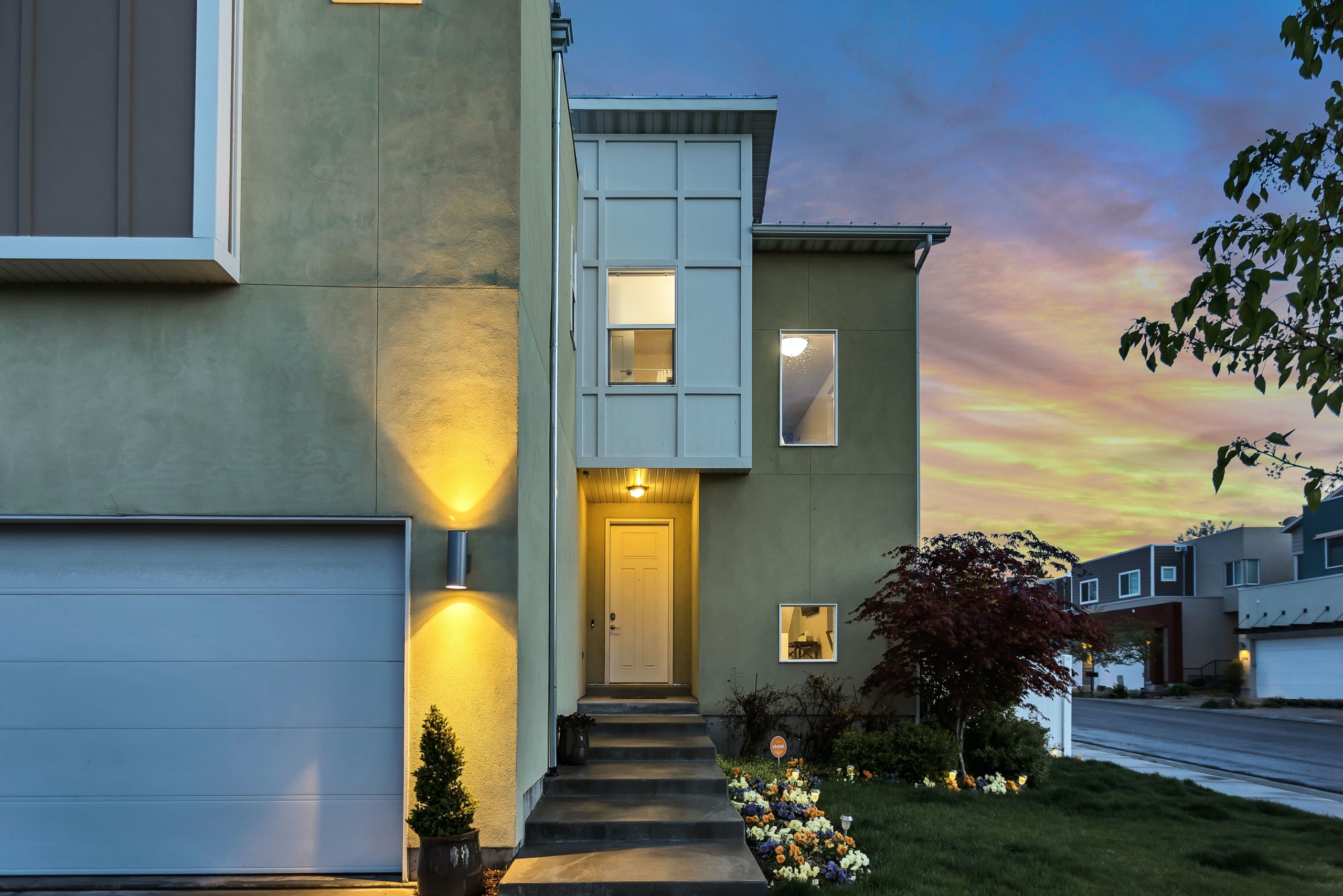 Image of modern townhouse with 1 car garage in front and cement pathway from garage/driveway to front door. At sunset with lights on. Two story with rectangle angles. Buying Your First Home across the image.