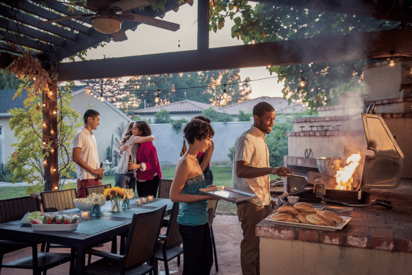 Photo of young adults in a wonderful backyard getting ready to eat under a back porch big enough for large dining table and large grill.  Entertaining backyards are one of the top home features in 2023