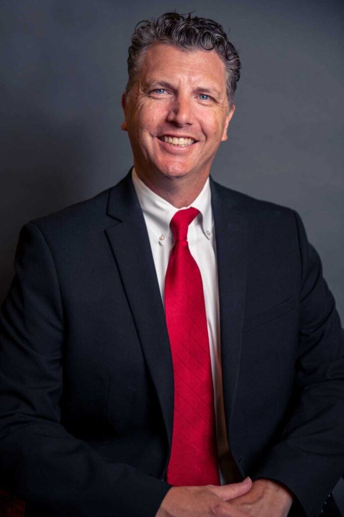 Head shot of Jay Hudson, Luxury Real Estate Agent, in black suite and red tie with grey background