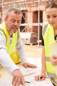 Two people in yellow caution vests looking a drawings on a build sight.  Older man with grey hair and younger female adult.  Both could be builders or one could be the builder and the other the buyer